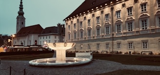 A large, historic building with ornate windows and a sloped roof stands prominently. In front of it is an illuminated fountain with a marble basin. The building is surrounded by a cobblestone pathway and a neatly kept lawn. Adjacent to the main building, there is a church with a tall steeple. The setting appears to be at dusk, with streetlights casting a warm glow.