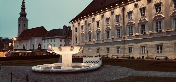 A large, historic building with ornate windows and a sloped roof stands prominently. In front of it is an illuminated fountain with a marble basin. The building is surrounded by a cobblestone pathway and a neatly kept lawn. Adjacent to the main building, there is a church with a tall steeple. The setting appears to be at dusk, with streetlights casting a warm glow.