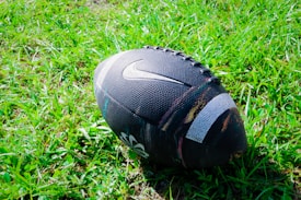 A black football with a textured surface is resting on a field of green grass. The ball features a prominent logo.