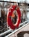 A bright orange lifebuoy is attached to a metallic railing, with some ropes hanging from it. The background features a blurred body of water and tall, dry reeds or grass. The lifebuoy has visible markings on it, including numbers and a label.