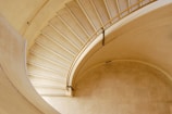A beautifully designed spiral staircase with a smooth, elegantly curving handrail made of metal. The staircase is crafted from light-colored stone, and the steps gently ascend, creating a sense of movement and flow. The overall structure is minimalist yet visually striking.
