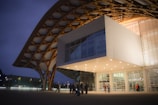 An architectural structure with a modern design, featuring a large wooden lattice roof that extends over a white cubic building. The building is illuminated with bright lights and contains large glass windows. People are gathered outside, creating a lively atmosphere. It's evening, and the sky is dimly lit.