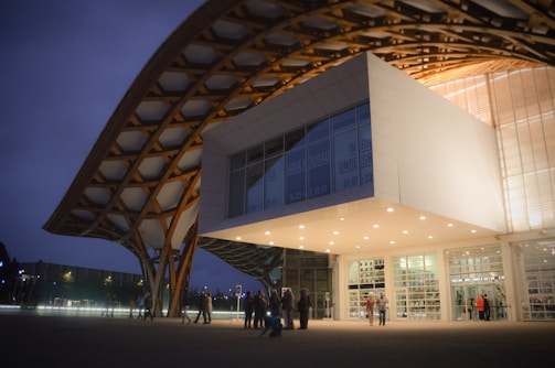 An architectural structure with a modern design, featuring a large wooden lattice roof that extends over a white cubic building. The building is illuminated with bright lights and contains large glass windows. People are gathered outside, creating a lively atmosphere. It's evening, and the sky is dimly lit.