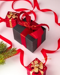 A collection of neatly wrapped gift boxes in various sizes, predominantly featuring red and black wrapping paired with gold accents and ribbons. A black box with a red ribbon is placed centrally, accompanied by smaller red boxes adorned with golden decorations. Bright red ribbon loops artistically across the background, and a sprig of evergreen adds a festive touch.