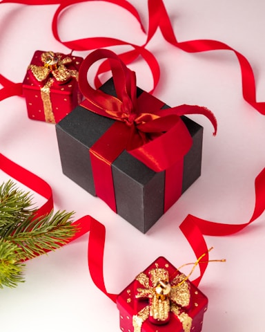 A collection of neatly wrapped gift boxes in various sizes, predominantly featuring red and black wrapping paired with gold accents and ribbons. A black box with a red ribbon is placed centrally, accompanied by smaller red boxes adorned with golden decorations. Bright red ribbon loops artistically across the background, and a sprig of evergreen adds a festive touch.