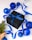 A black gift box adorned with a blue ribbon is surrounded by shiny blue Christmas ornaments on a white surface. A blue ribbon is elegantly draped around the items, which are accompanied by a small sprig of evergreen wrapped in burlap.