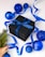 A black gift box adorned with a blue ribbon is surrounded by shiny blue Christmas ornaments on a white surface. A blue ribbon is elegantly draped around the items, which are accompanied by a small sprig of evergreen wrapped in burlap.