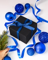 A black gift box adorned with a blue ribbon is surrounded by shiny blue Christmas ornaments on a white surface. A blue ribbon is elegantly draped around the items, which are accompanied by a small sprig of evergreen wrapped in burlap.