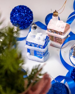 Festive decorations featuring shiny, miniature house ornaments in blue and pink with glitter. Surrounding them are vibrant blue ribbons, sparkly spherical ornaments, and a hint of green foliage. The setting is bright, adding a cheerful, holiday vibe.