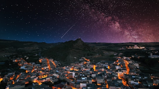 A small town illuminated by street lights during nighttime, nestled in a valley beneath a dark sky filled with stars. The Milky Way is visible, creating a stunning backdrop over the silhouette of a mountain.