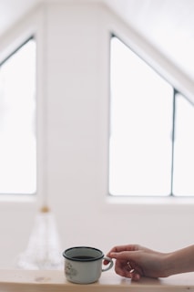 A close-up of a hand holding a delicate enamel pin shaped like a steaming coffee cup, resting on a soft linen background.