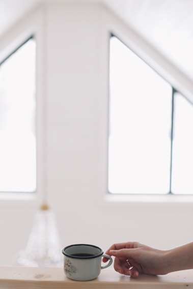 A close-up of a hand holding a delicate enamel pin shaped like a steaming coffee cup, resting on a soft linen background.
