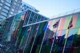 A modern glass building with colorful, reflective panels is showcased. Several international flags are displayed along the length of the building. Adjacent to it, a high-rise structure is also visible. The sky is clear and blue.