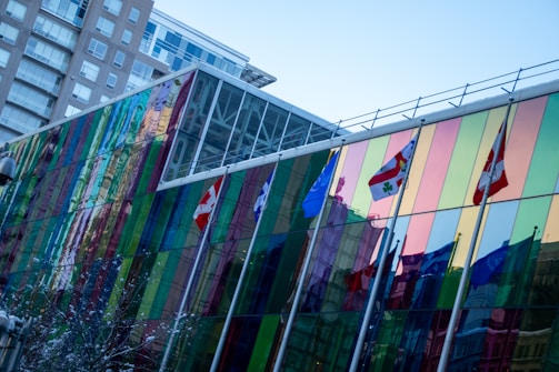 A modern glass building with colorful, reflective panels is showcased. Several international flags are displayed along the length of the building. Adjacent to it, a high-rise structure is also visible. The sky is clear and blue.