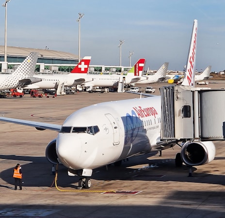 A busy airport scene featuring multiple airplanes parked at the gates. A prominent aircraft with an Air Europa logo is connected to a jet bridge. Several other planes are visible in the background, each with distinct tail designs. An airport worker wearing an orange vest stands near the Air Europa plane.