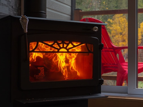 A wood-burning stove radiates warmth with visible orange flames behind a decorative grate. Next to the stove, a large window reveals an outdoor scene with red Adirondack chairs and autumnal trees with yellow leaves.