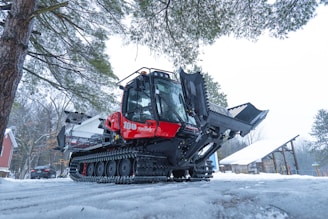 A large snow grooming machine with caterpillar tracks is parked on a snowy path surrounded by trees. The machine has a bright red cabin and black grooming equipment. There is snow on the ground and a building with a peaked roof in the background.