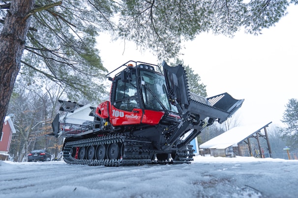 A large snow grooming machine with caterpillar tracks is parked on a snowy path surrounded by trees. The machine has a bright red cabin and black grooming equipment. There is snow on the ground and a building with a peaked roof in the background.