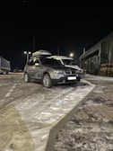 A silver SUV is parked on a snowy road at night. The vehicle has a roof box attached. Streetlights illuminate the area, casting shadows on the snow-covered ground. There is a truck parked nearby, and an industrial building with stacked wooden pallets can be seen in the background.