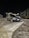 A silver SUV is parked on a snowy road at night. The vehicle has a roof box attached. Streetlights illuminate the area, casting shadows on the snow-covered ground. There is a truck parked nearby, and an industrial building with stacked wooden pallets can be seen in the background.