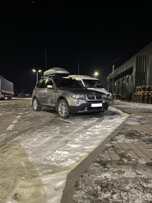 A silver SUV is parked on a snowy road at night. The vehicle has a roof box attached. Streetlights illuminate the area, casting shadows on the snow-covered ground. There is a truck parked nearby, and an industrial building with stacked wooden pallets can be seen in the background.
