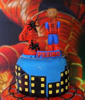 A colorful cake designed with a superhero theme featuring a blue and red color scheme. The top features a small figure in a superhero costume with a number four candle that includes spider decorations. The base of the cake is decorated with a cityscape design.