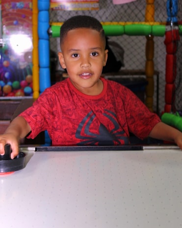 A young child with short hair is playing air hockey, wearing a red Spider-Man themed shirt. The background features colorful play equipment, including bright tubes and a netted structure.