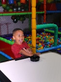A child with an excited expression plays air hockey in a colorful play area filled with plastic balls. The environment is vibrant with structures made of padded bars in various colors.