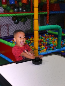 A child with an excited expression plays air hockey in a colorful play area filled with plastic balls. The environment is vibrant with structures made of padded bars in various colors.