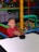 A child with an excited expression plays air hockey in a colorful play area filled with plastic balls. The environment is vibrant with structures made of padded bars in various colors.