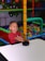 A child with an excited expression plays air hockey in a colorful play area filled with plastic balls. The environment is vibrant with structures made of padded bars in various colors.