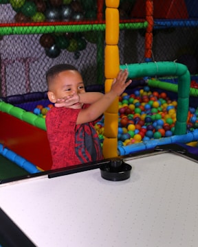A young boy wearing a red shirt is at an indoor play area. He appears to be having fun as he poses near a ball pit filled with colorful plastic balls. The play area is enclosed by a net and features bright, multi-colored foam padding.