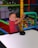 A young boy wearing a red shirt is at an indoor play area. He appears to be having fun as he poses near a ball pit filled with colorful plastic balls. The play area is enclosed by a net and features bright, multi-colored foam padding.