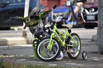 Two children's bicycles are parked and secured with a lock on a city sidewalk. One of the bikes is lime green with training wheels, while the other has a distinctive black handlebar shield with text. The background includes parked cars and a blurred figure, indicating an urban setting.