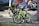 Two children's bicycles are parked and secured with a lock on a city sidewalk. One of the bikes is lime green with training wheels, while the other has a distinctive black handlebar shield with text. The background includes parked cars and a blurred figure, indicating an urban setting.