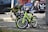 Two children's bicycles are parked and secured with a lock on a city sidewalk. One of the bikes is lime green with training wheels, while the other has a distinctive black handlebar shield with text. The background includes parked cars and a blurred figure, indicating an urban setting.