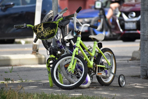 Two children's bicycles are parked and secured with a lock on a city sidewalk. One of the bikes is lime green with training wheels, while the other has a distinctive black handlebar shield with text. The background includes parked cars and a blurred figure, indicating an urban setting.