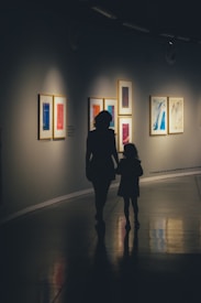 Silhouettes of an adult and a child walking through a dimly lit art gallery. Framed colorful artworks hang on the walls, creating a contrast against the muted lighting. The polished floor reflects their shadows softly, emphasizing the serene ambiance.