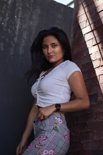 A woman with long, dark hair is wearing a white, short-sleeved top and patterned pants featuring floral designs. She stands against a backdrop of textured, brick and concrete walls, partially illuminated by natural light from above.