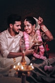 A couple is sitting closely together, dressed in traditional attire. The woman is wearing a heavily embroidered blouse with an elaborate pink garland and artistic henna on her hands. She holds a traditional oil lamp, and both are lovingly gazing at each other, creating an intimate and cultural atmosphere.