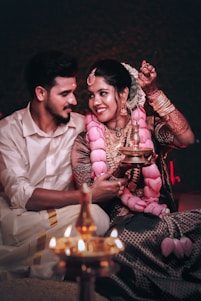A couple is sitting closely together, dressed in traditional attire. The woman is wearing a heavily embroidered blouse with an elaborate pink garland and artistic henna on her hands. She holds a traditional oil lamp, and both are lovingly gazing at each other, creating an intimate and cultural atmosphere.
