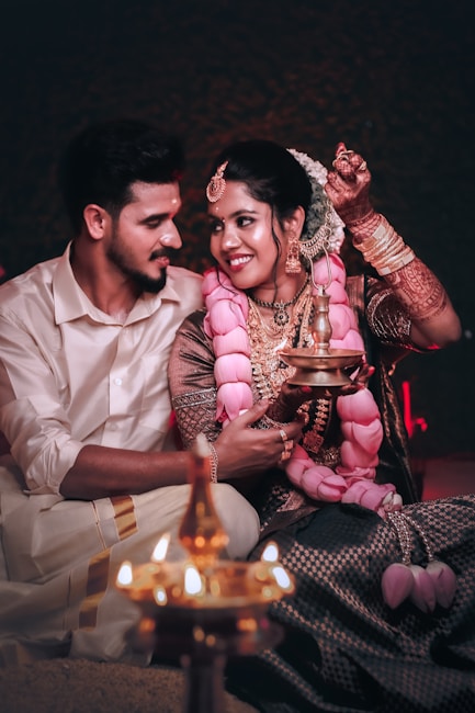 A couple is sitting closely together, dressed in traditional attire. The woman is wearing a heavily embroidered blouse with an elaborate pink garland and artistic henna on her hands. She holds a traditional oil lamp, and both are lovingly gazing at each other, creating an intimate and cultural atmosphere.