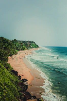 A serene beach with people leisurely walking and enjoying the shoreline. The coastline is bordered by lush green vegetation and cliffs on one side, and the blue-green ocean on the other. The sandy beach extends into the distance with gentle waves lapping against the shore.