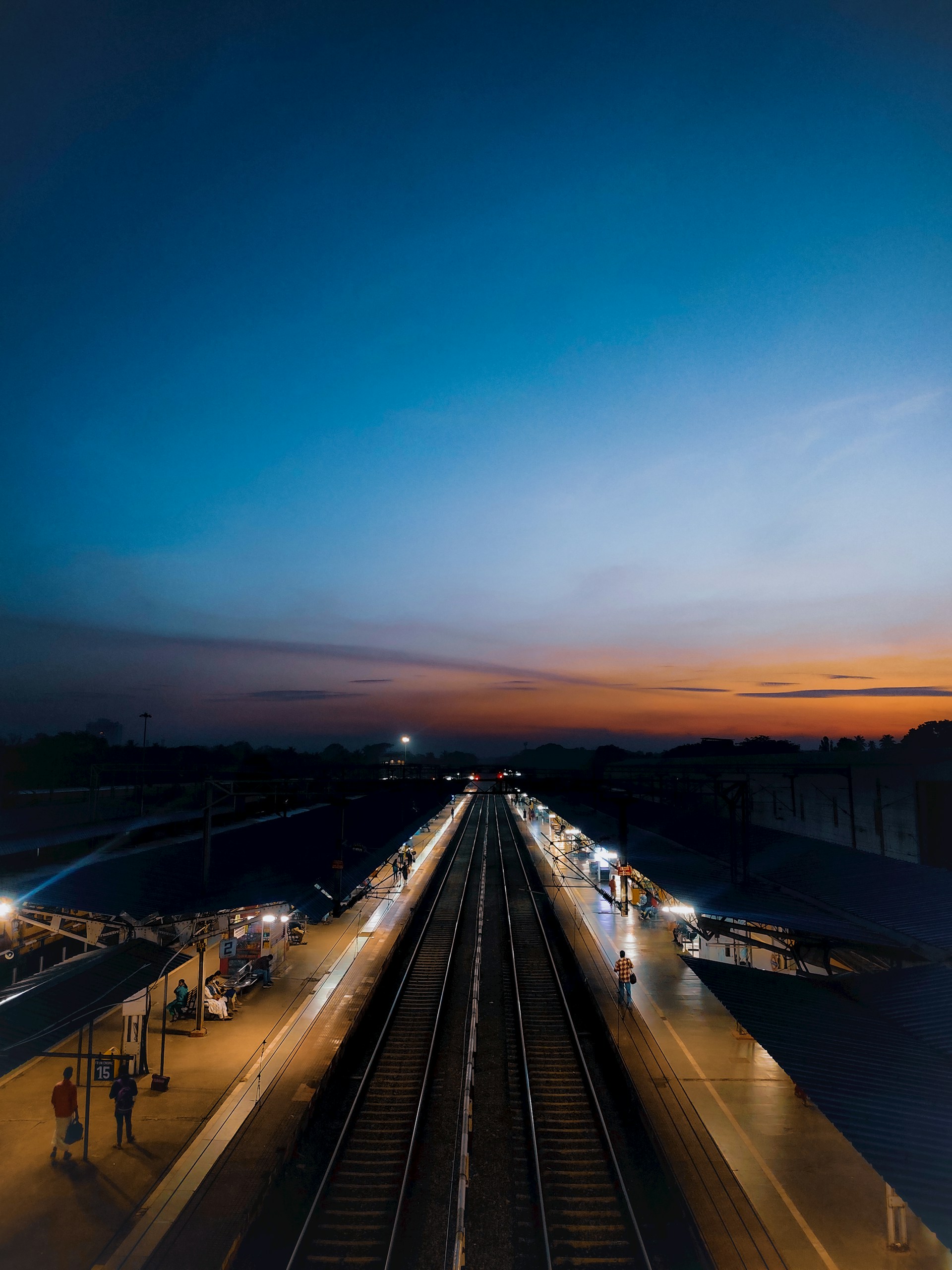 A train platform stretches into the distance under a twilight sky. The platform is lined with lights and small groups of people. The sky transitions from deep blue at the top to warm oranges and pinks near the horizon.