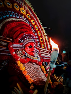 A vibrant traditional costume featuring an elaborate headdress adorned with intricate patterns and bright colors, predominantly red and yellow. The costume includes a striking face mask with detailed designs and a torch adds dramatic lighting to the scene, casting a warm glow.