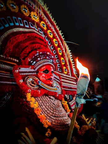 A vibrant traditional costume featuring an elaborate headdress adorned with intricate patterns and bright colors, predominantly red and yellow. The costume includes a striking face mask with detailed designs and a torch adds dramatic lighting to the scene, casting a warm glow.