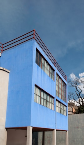 A modern architectural structure with a vibrant blue exterior stands prominently. The building features large glass windows and red railings along the top level. The sky is clear, accentuating the bold colors of the structure.