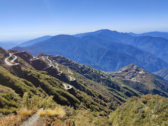 A winding mountain road cutting through lush green hills under a bright blue sky.