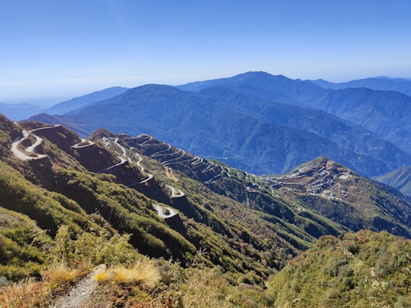 A scenic mountain road winding through lush green hills under a clear blue sky.
