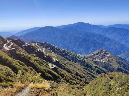 A winding mountain road cutting through lush green hills under a bright blue sky.